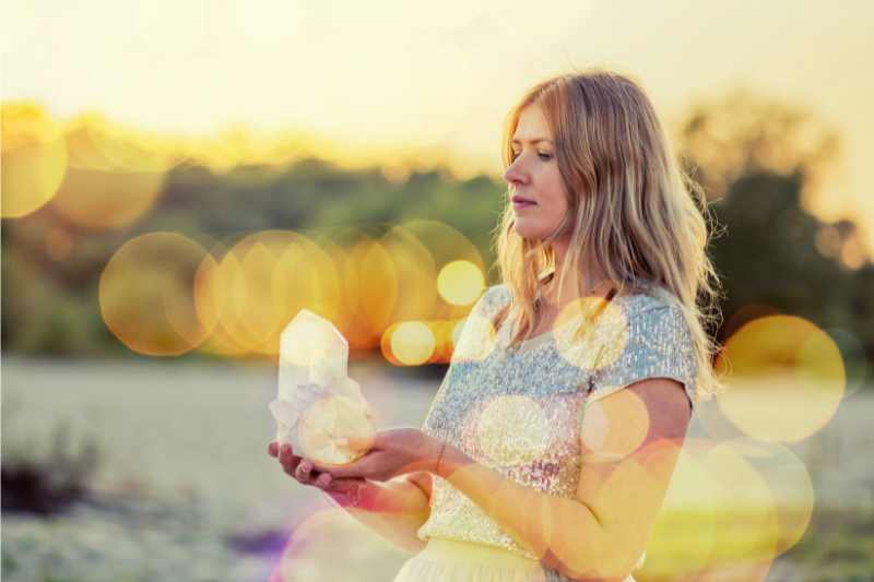 Eine Frau mit blonden langen Haaren im Abendlicht mit goldenen Lichtreflexen über das Bild verteilt hält einen Bergkristall in den Händen.