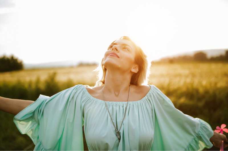 Eine glückliche Frau mit weißter Bluse vor dem Hintergrund eines gelben Feldes in Abendlicht breitet die Arme aus Eine glückliche Frau mit einem Getreidefeld und weiter Landschaft im Hintergrund, gekleidet mit einer weißen Trachtenbluse im Balkanstyle mit weiten Ärmeln und mit langer Halskette breiten breitet ihre Arme aus und schaut mit einem Gefühl der Freiheit in den Himmel